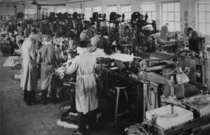 Blick in die historische Produktionshalle in Triptis mit Mitarbeitenden an Maschinenarbeitsplätzen in Arbeitskitteln.