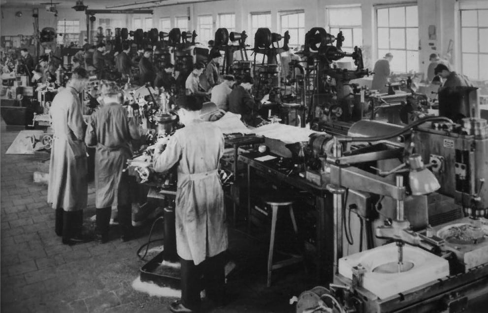 Blick in die historische Produktionshalle in Triptis mit Mitarbeitenden an Maschinenarbeitsplätzen in Arbeitskitteln.